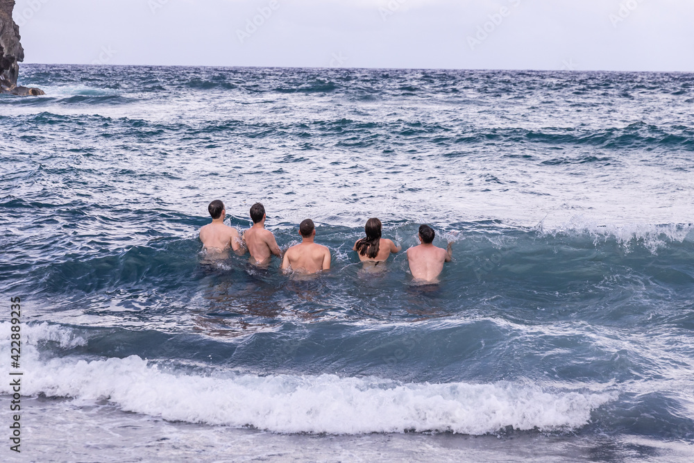 Group of young people - one girl and four boys jump into the incoming wave during high tide on a Waianapanapa black beach, Maui, Hawaii