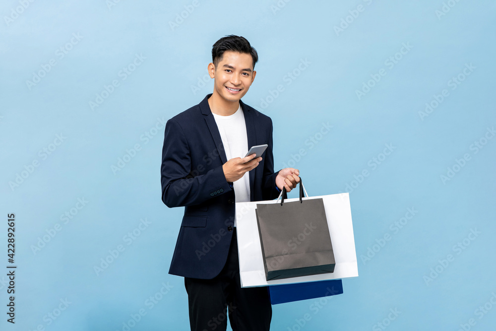 Young handsome southeast Asian man holding mobile phone and shopping bags in light blue studio isolated background Young handsome southeast Asian man holding mobile phone and shopping bags in light blue studio isolated background