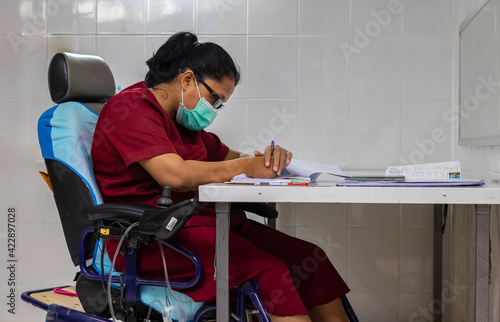 A woman with Quadriplegia sitting in her electric wheelchair while using her both hands doing paperwork on the desk in her office. photo