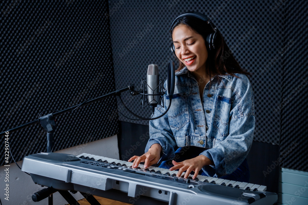 Happy cheerful pretty smiling of young Asian woman vocalist Wearing Headphones recording a song front of microphone and playing the keyboard during rehearsal of her band in a professional studio