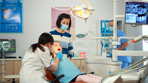 Dentist technician with gloves explaining the process to kid patient before surgery lighting the lamp. Orthodontist speaking to girl with toothache sitting on stomatological chair.