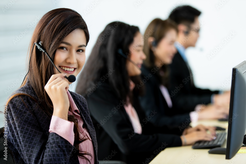 Cheerful Asian woman in headset smile with cheerful and happy while working on computer at desk with coworkers in help desk office. Sevice mind and ready for marketing support concept. Cheerful Asian woman in headset smile with cheerful and happy while working on computer at desk with coworkers in help desk office. Sevice mind and ready for marketing support concept.