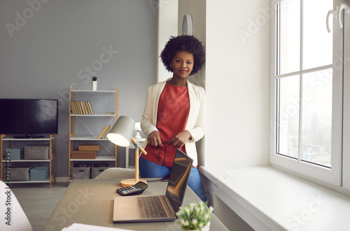 Young successful african american businesswoman in office portrait. Smiling black woman entrepreneur standing next to window rest after hard work looking at camera. Self-confidence and success photo