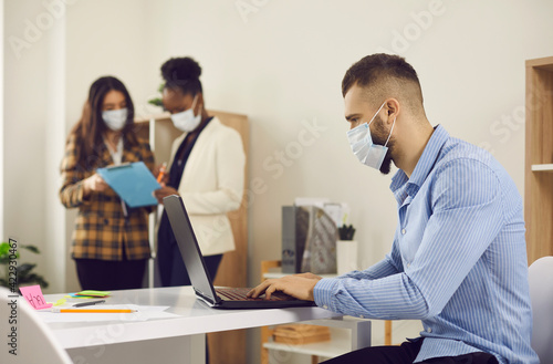 Man employee in face medical mask working on laptop indoors in office after lockdown with interracial colleague on background. New normal business practice of coronavirus covid-19 outbreak control