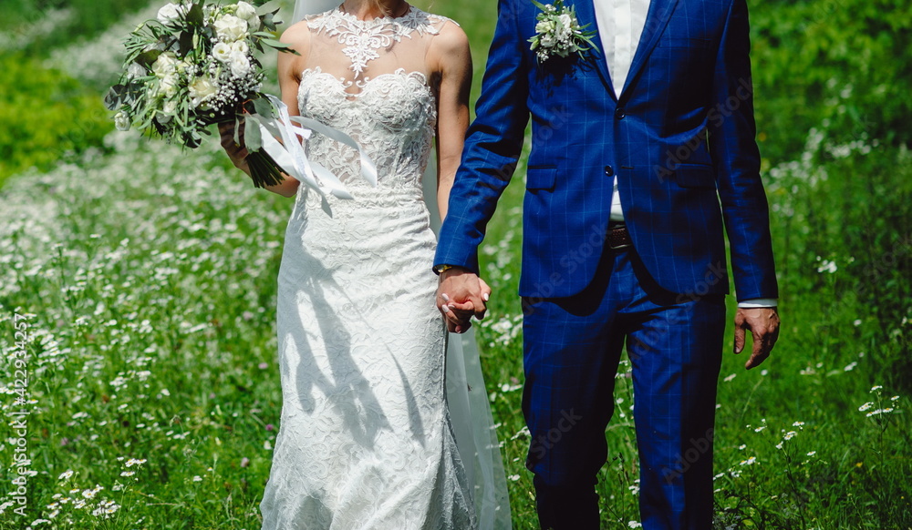 Bride in white dress with lace and groom in tuxedo hold each other hands on a background of green grass and flowers. Wedding ceremony outdoors. Bride in white dress with lace and groom in tuxedo hold each other hands on a background of green grass and flowers. Wedding ceremony outdoors.