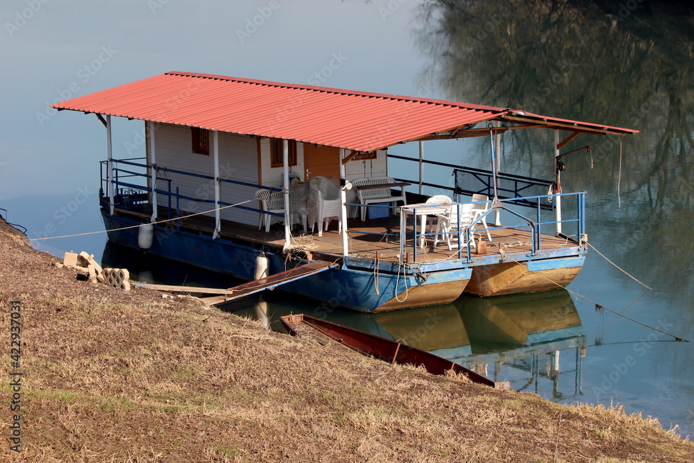 River barge converted into river boat house with improvised homemade roof and terrace with white plastic chairs and tables left at local river bank covered with dry grass on cold sunny winter day