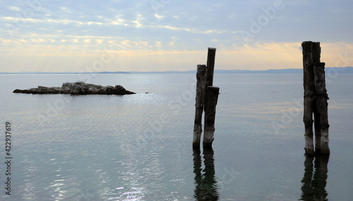 Wooden poles and a rock emerge from the water. Peace and silence on Lake Garda. The sun filters through the clouds before sunset.