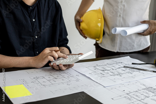 The architect was pressing the white calculator and there was an engineer standing there holding the floor plan. They are examining the draft blueprint for the designed building. Construction ideas.