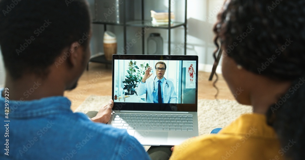 Rear of African American couple man and woman sitting in living room in house and videochatting on laptop on online video call with young handsome male doctor in white coat, consultation, close up Rear of African American couple man and woman sitting in living room in house and videochatting on laptop on online video call with young handsome male doctor in white coat, consultation, close up