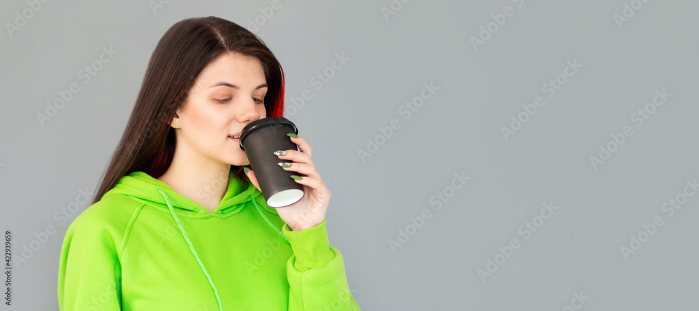Studio shot of girl with holds paper cup of hot coffee, wears green jumper, grey background. Beautiful young woman enjoys aromatic cappuccino. Studio shot of girl with holds paper cup of hot coffee, wears green jumper, grey background. Beautiful young woman enjoys aromatic cappuccino.