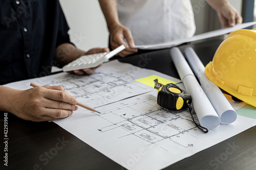 The architect was pressing the white calculator and there was an engineer standing there holding the floor plan. They are examining the draft blueprint for the designed building.