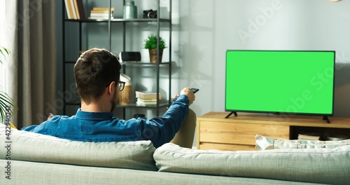 Fototapeta Rear of Caucasian young guy watching TV with chroma key resting on sofa sitting in living room in house