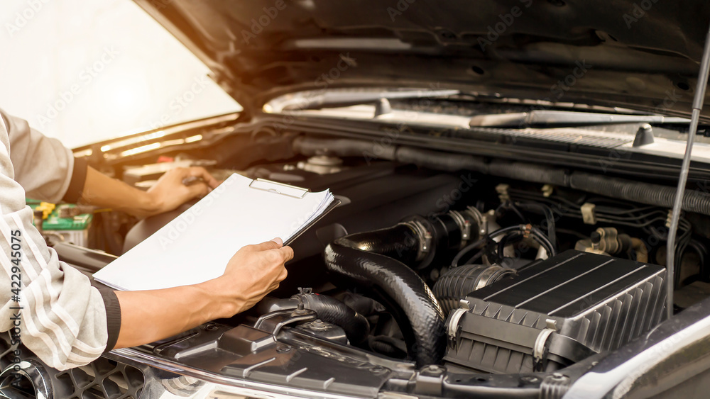 Automobile mechanic repairman checking a car engine with inspecting writing to the clipboard the checklist for repair machine, car service and maintenance. Automobile mechanic repairman checking a car engine with inspecting writing to the clipboard the checklist for repair machine, car service and maintenance.