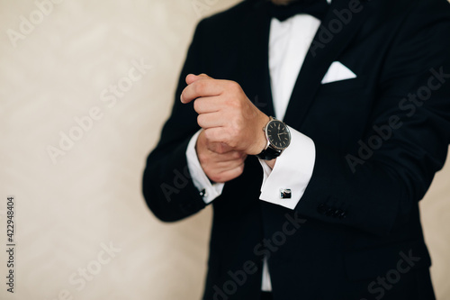 Businessman adjusts his white shirt button. Concept successful businessman. Portrait of trendy attractive stunning man in black tuxedo with tie fasten button on sleeve cuffs of his white shirt.