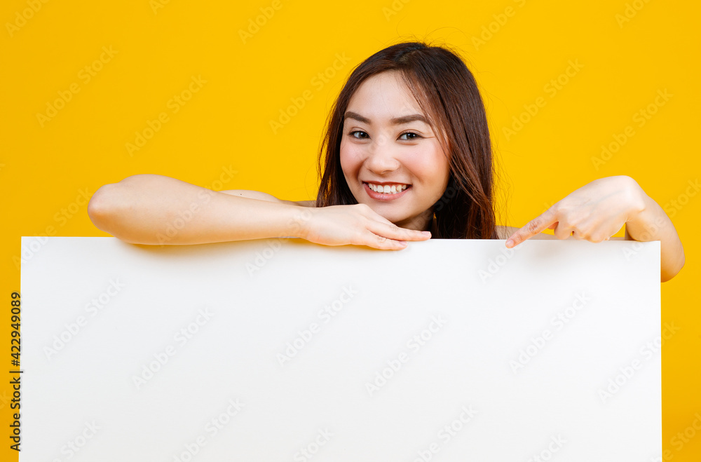 Cute and pretty curly hair Asian female brunette holding and pointing at white blank board poses to camera with a joyful for advertising and banner use purpose, isolated on bright yellow background Cute and pretty curly hair Asian female brunette holding and pointing at white blank board poses to camera with a joyful for advertising and banner use purpose, isolated on bright yellow background