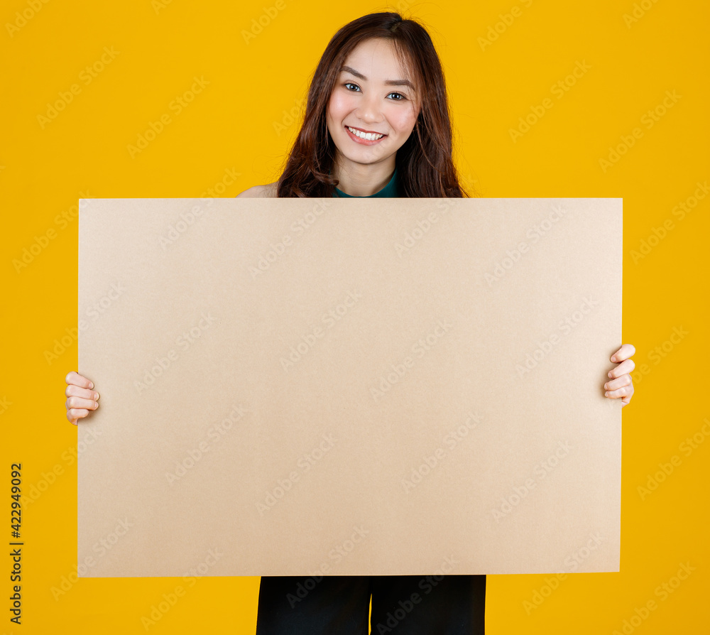 Cute and pretty curly hair Asian female brunette holding brown blank board poses to camera with a joyful for advertising and banner use purpose, studio shot isolated on bright yellow background Cute and pretty curly hair Asian female brunette holding brown blank board poses to camera with a joyful for advertising and banner use purpose, studio shot isolated on bright yellow background