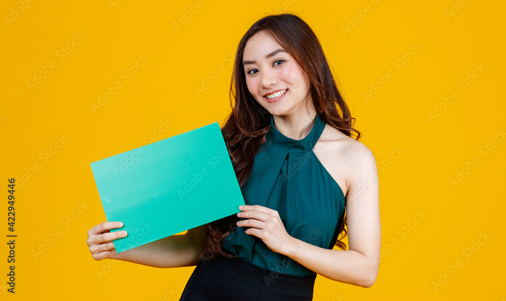Cute and pretty curly hair Asian female brunette holding green blank board poses to camera with a joyful and positive gesture for advertising use purpose, studio shot isolated on yellow background. Cute and pretty curly hair Asian female brunette holding green blank board poses to camera with a joyful and positive gesture for advertising use purpose, studio shot isolated on yellow background.
