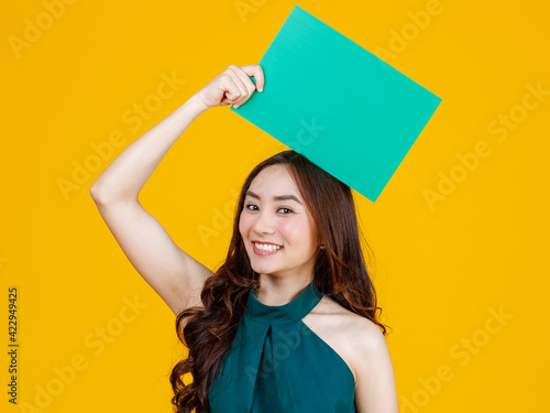Cute and pretty curly hair Asian female brunette holding green blank board poses to camera with a joyful and positive gesture for advertising use purpose, studio shot isolated on yellow background.