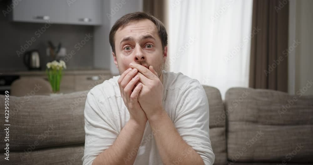 Shocked man looking at camera. Astonished adult male covering mouth and looking at camera in disbelief while sitting on couch at home Shocked man looking at camera. Astonished adult male covering mouth and looking at camera in disbelief while sitting on couch at home