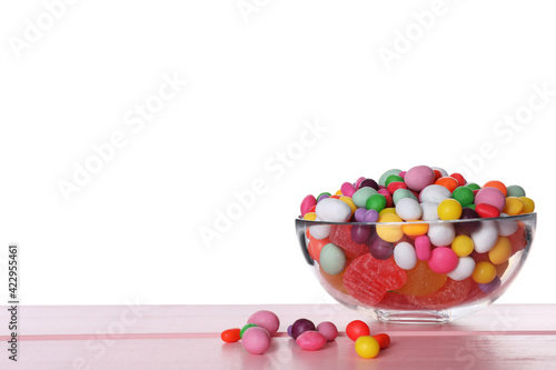 Many different candies in glass bowl on pink wooden table against white background