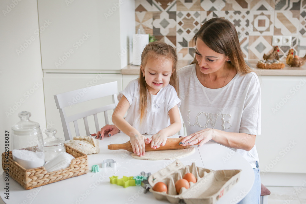 Happy mom and daughter prepare dough together using rolling pin, smiling young mom, charming girl prepare cookies, buns in the kitchen. The concept of a friendly family preparing for Easter Happy mom and daughter prepare dough together using rolling pin, smiling young mom, charming girl prepare cookies, buns in the kitchen. The concept of a friendly family preparing for Easter