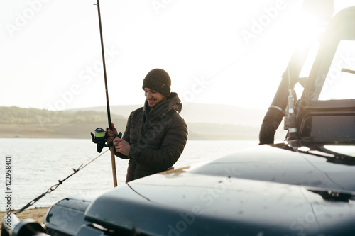 Young fisherman standing near his car and holding fishing equipment