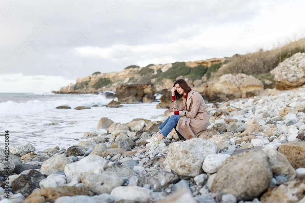 A beautiful young woman sits on a rocky shore and thinks about something. Evening portrait of a beautiful young woman in nature. Solitude, retreat, isoloation, seclusion concept