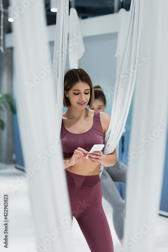 Young sportswoman using smartphone near fly yoga straps on blurred foreground