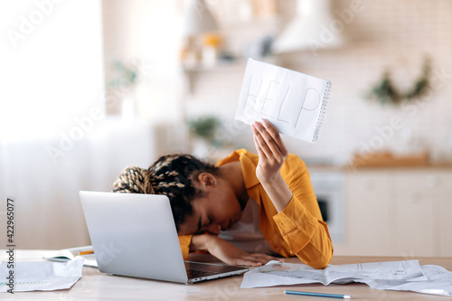 A frustrated African American female student is tired of studying at home, lies at her desk and holds a leaflet with the inscription help. Freelancer young woman need a rest from remotely work photo