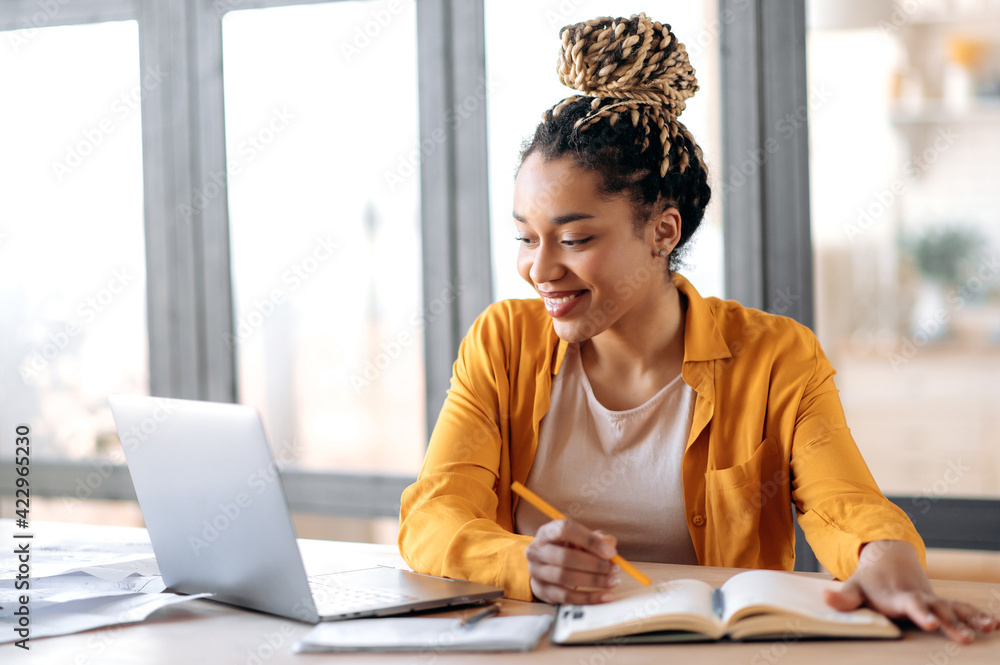 Online education, e-learning. Happy african american young woman in stylish casual clothes, studying remotely, using a laptop, listening to online lecture, taking notes while sitting at home, smiles - obrazy, fototapety, plakaty 