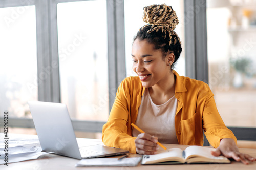 Online education, e-learning. Happy african american young woman in stylish casual clothes, studying remotely, using a laptop, listening to online lecture, taking notes while sitting at home, smiles