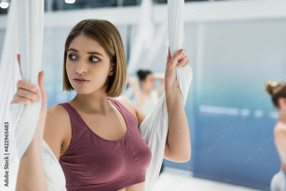sportive woman looking away while stretching with aerial yoga strap on blurred background