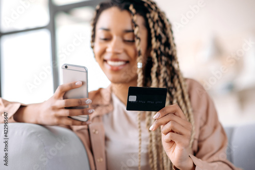 Defocused african american joyful smiling girl holding smartphone and bank card, shopping online, paying bills or ordering home delivery, entering card number, e-payment concept