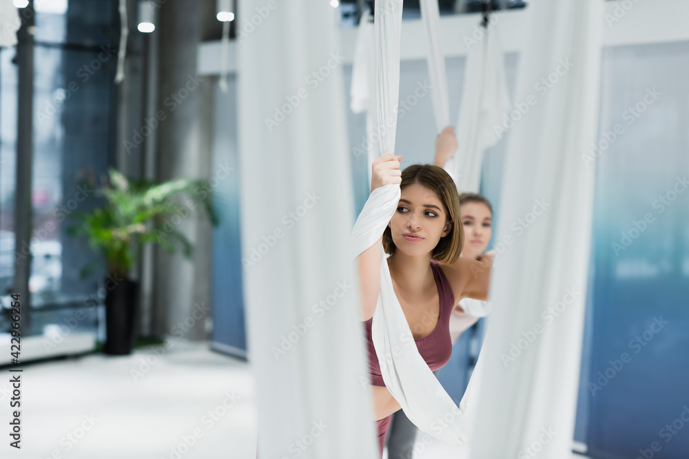 young woman looking away while stretching with fly yoga hammock on blurred foreground