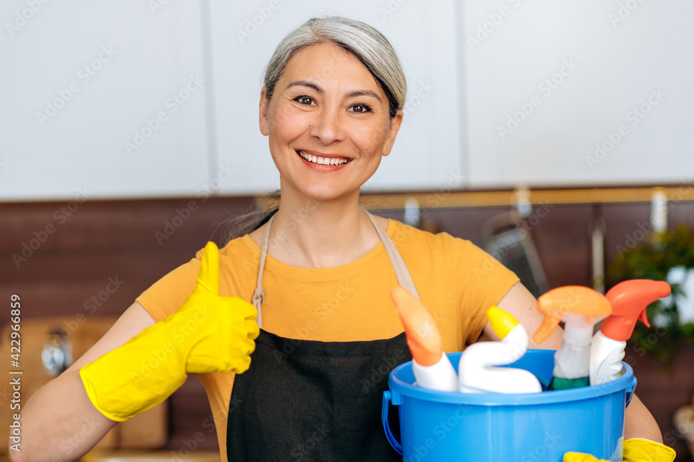 Cleaning service concept. Portrait of happy senior gray-haired Asian cleaning lady or housewife, wearing apron and gloves, holding bucket, shows thumb up, looks at camera, smiling, ready for cleaning Cleaning service concept. Portrait of happy senior gray-haired Asian cleaning lady or housewife, wearing apron and gloves, holding bucket, shows thumb up, looks at camera, smiling, ready for cleaning