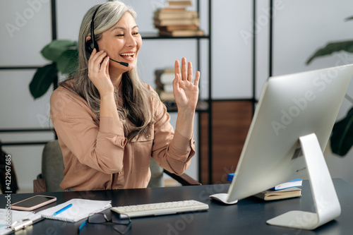 Successful confident senior Asian gray-haired female business lady, manager, coach, wearing headset, chats with business partners by conference call, online briefing, gesturing hands, friendly smile