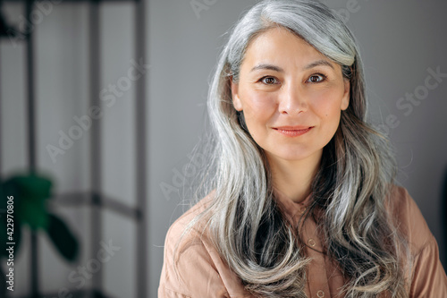Close-up portrait of beautiful influential confident mature gray-haired Asian woman ceo, lawyer, real estate agent, stands in the office, dressed in stylish clothes, looks at camera, friendly smiling