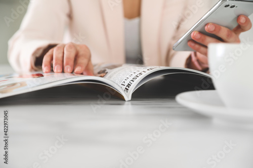 Фотография Woman with mobile phone reading magazine at white table, closeup