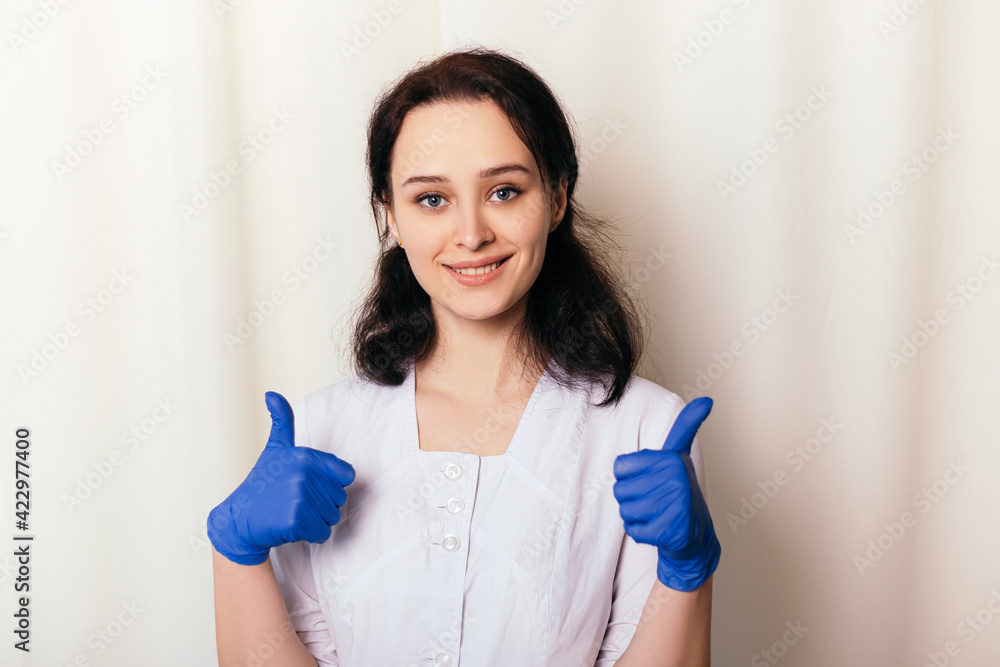 Nurse young medic rubber disinfected gloves show like thumbs up. Female doctor dressed in medical gown happy smiling cheerful. Copy space. Pandemic virus coronavirus concept Nurse young medic rubber disinfected gloves show like thumbs up. Female doctor dressed in medical gown happy smiling cheerful. Copy space. Pandemic virus coronavirus concept