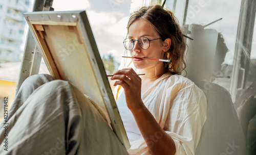 Closeup portrait of a pretty female artist painting on canvas in her art studio sitting next to the window. A woman painter with eyeglasses painting with oil searching for imagination in the workshop. photo