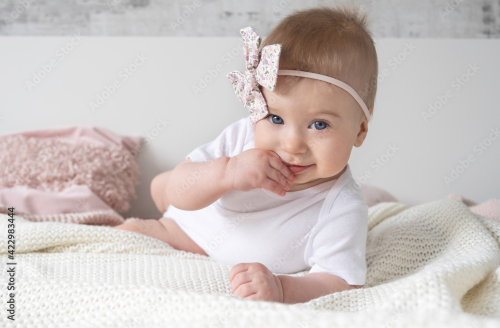 Beaytiful caucasian blonde baby girl with pink bow on head lying ob bed, putting finger into mouth looking at camera on background at home.Family lifestyle,kid fashion accessories Beaytiful caucasian blonde baby girl with pink bow on head lying ob bed, putting finger into mouth looking at camera on background at home.Family lifestyle,kid fashion accessories