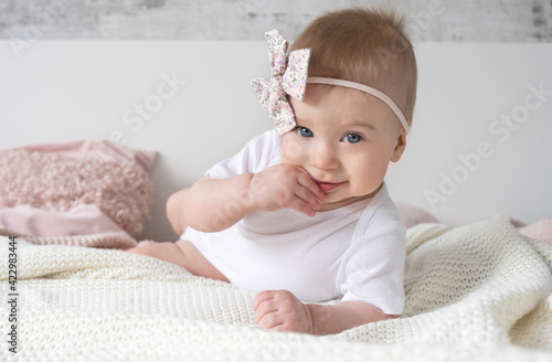 Beaytiful caucasian blonde baby girl with pink bow on head lying ob bed, putting finger into mouth looking at camera on background at home.Family lifestyle,kid fashion accessories