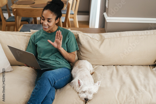 Black woman gesturing and using laptop while sitting with her dog on sofa