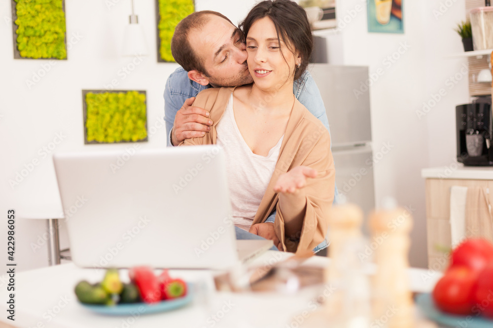Husband kissing wife cheek while reading ideas for breakfast in kitchen. Happy loving cheerful romantic in love couple at home using modern wifi wireless internet technology Husband kissing wife cheek while reading ideas for breakfast in kitchen. Happy loving cheerful romantic in love couple at home using modern wifi wireless internet technology