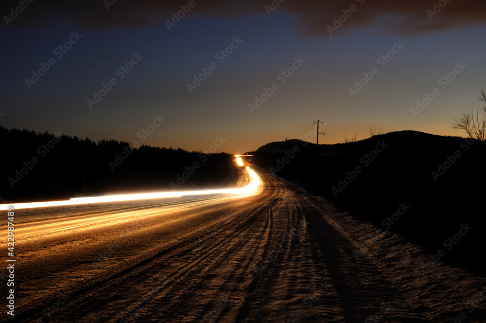 An asphalt road at dusk with the headlights of passing cars at a slow shutter speed. Night view with long passing headlights. Wallpaper.