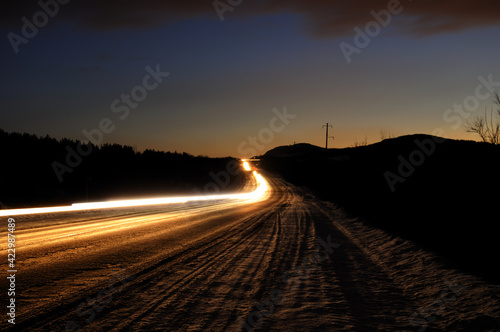 An asphalt road at dusk with the headlights of passing cars at a slow shutter speed. Night view with long passing headlights. Wallpaper.