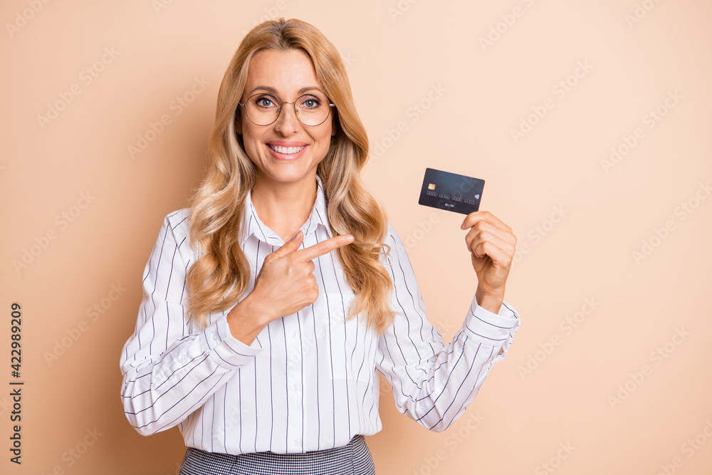 Photo of adorable sweet mature woman dressed white shirt glasses holding pointing bank card isolated beige color background Photo of adorable sweet mature woman dressed white shirt glasses holding pointing bank card isolated beige color background