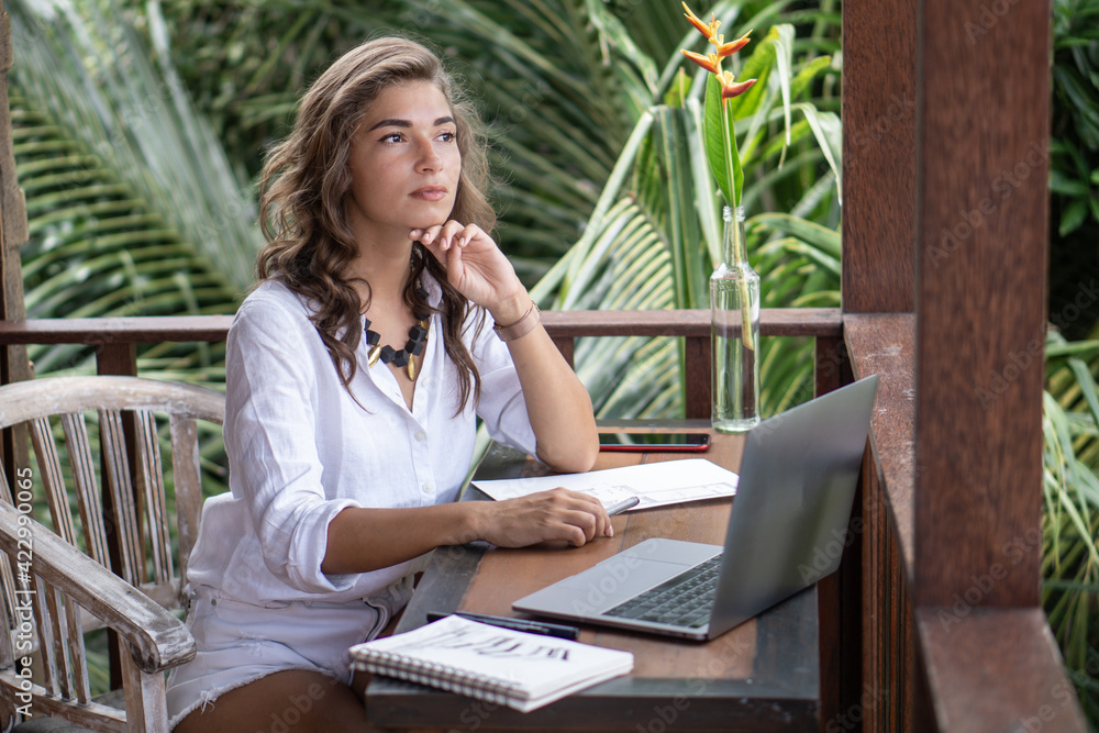 Portrait of young beautiful woman freelancer working with notebook, laptop in white shirt on balcony of tropical bungalow with palm trees view Portrait of young beautiful woman freelancer working with notebook, laptop in white shirt on balcony of tropical bungalow with palm trees view