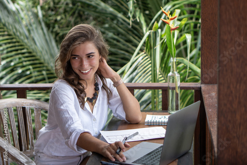 Portrait of young beautiful woman freelancer working with notebook, laptop in white shirt on balcony of tropical bungalow with palm trees view