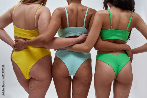 Back view of three multiethnic women with different body shapes in colorful underwear embracing each other while posing in the studio over light background